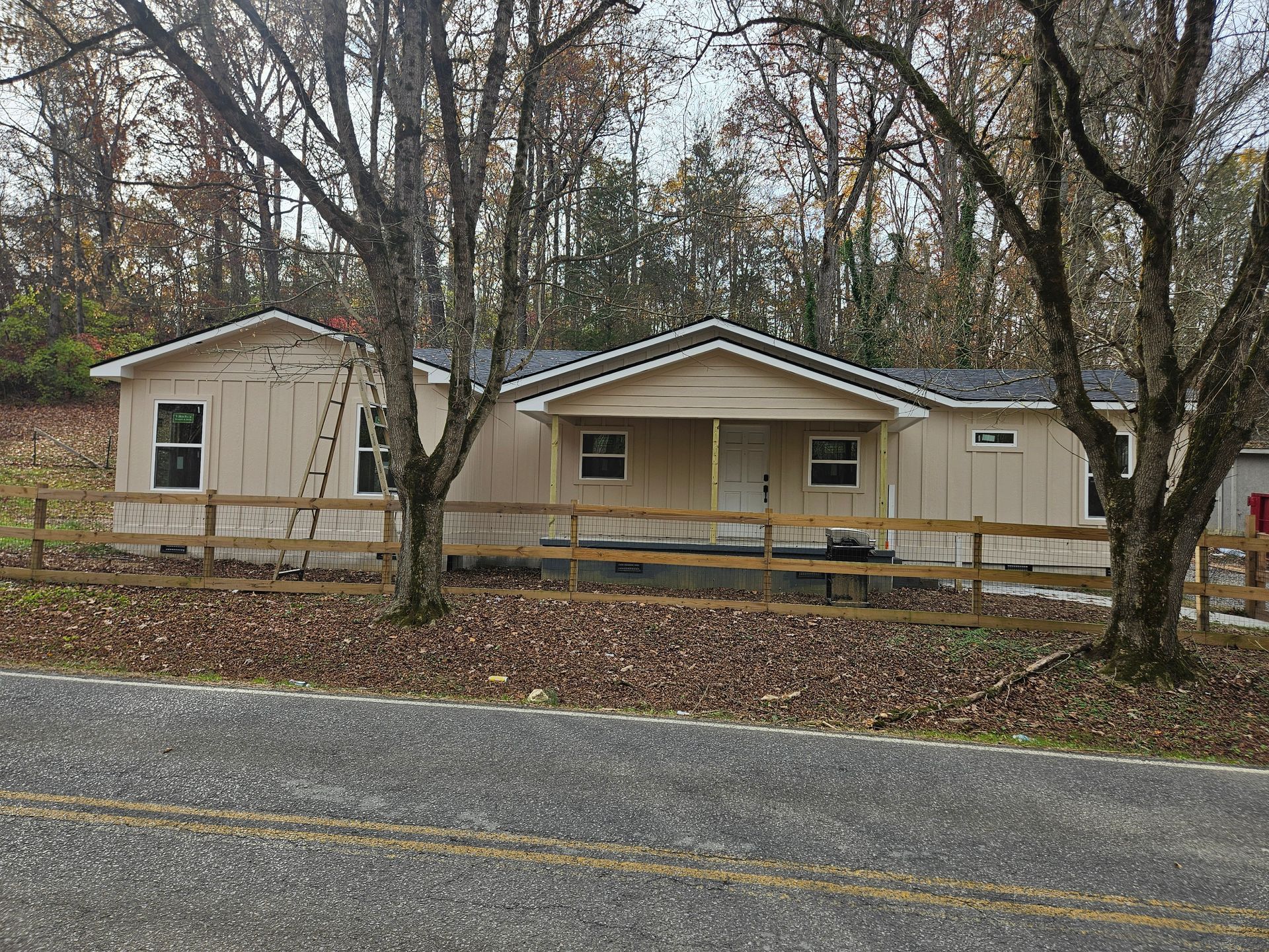 Tan single-story house with a small porch, wooden fence, and ladder; set among trees on a roadside.