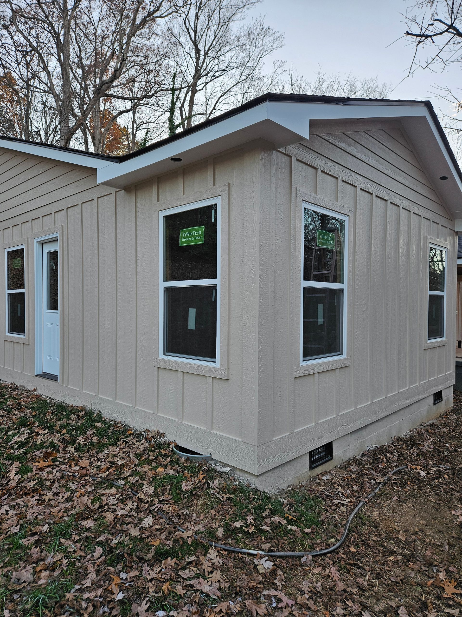 Beige-sided house with white trim and windows. Door visible. Dry leaves on the ground.