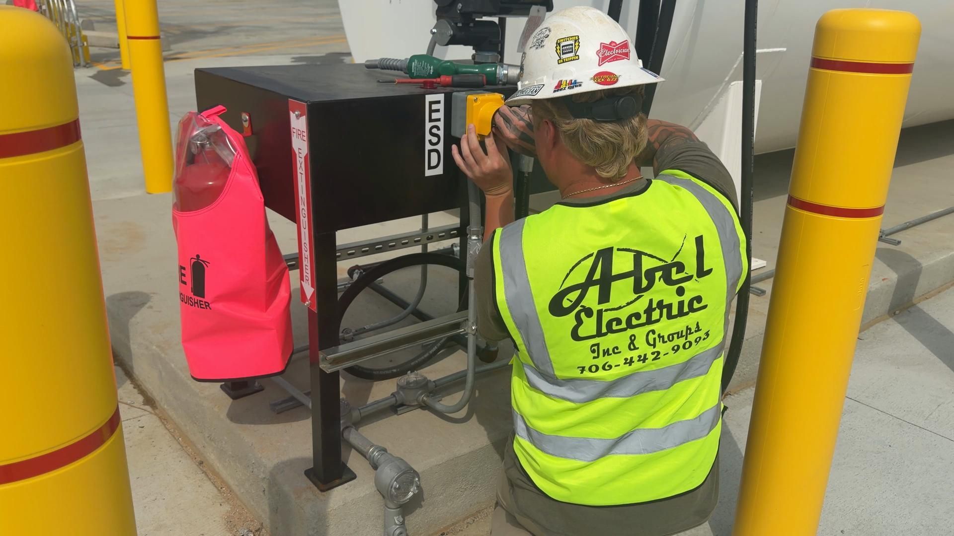 Worker in neon vest and hard hat using a power tool on equipment beside yellow safety bollards.