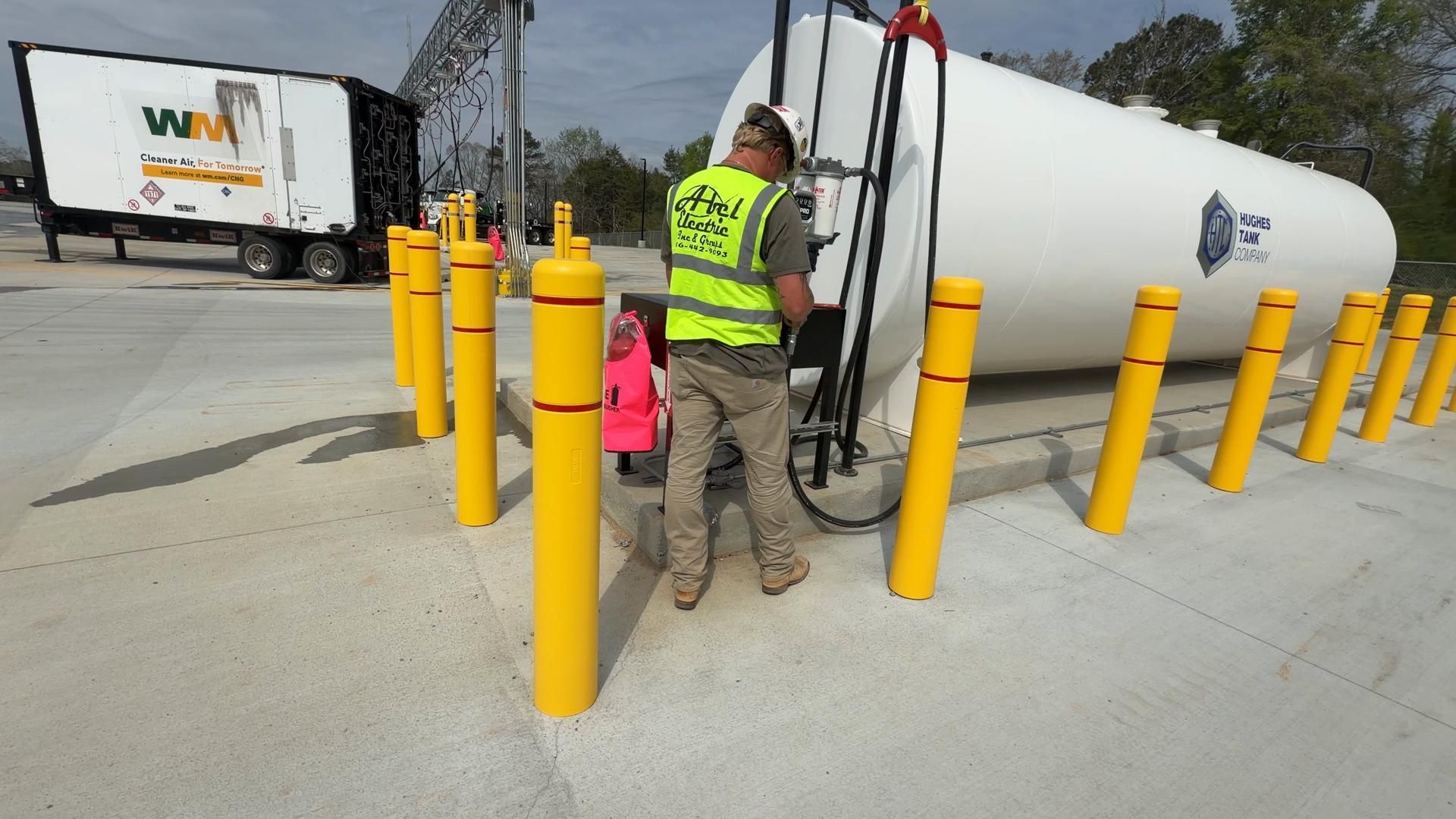 Workers in safety vests near a large white tank beside yellow bollards at an industrial site