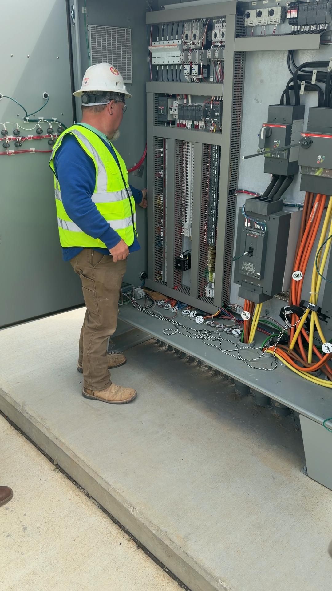 Worker in hard hat and safety vest inspecting an open electrical control cabinet outdoors.