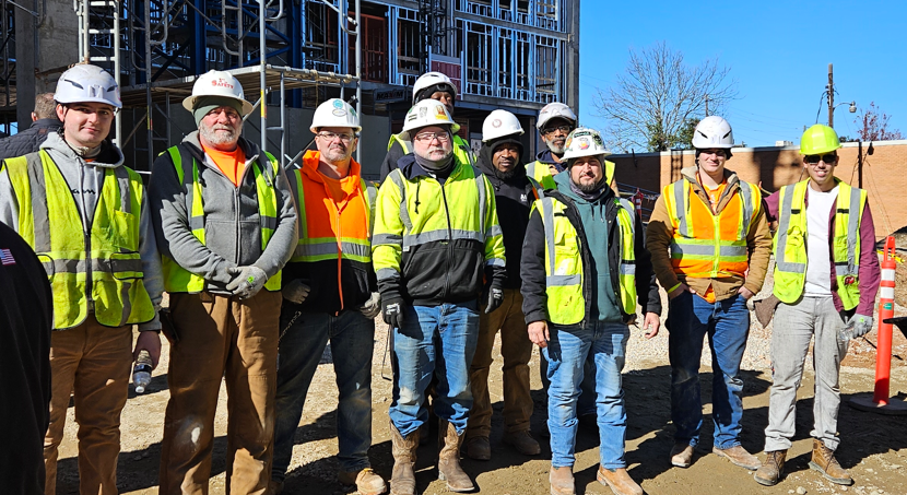 Construction crew in hard hats and neon safety vests standing at a building site.