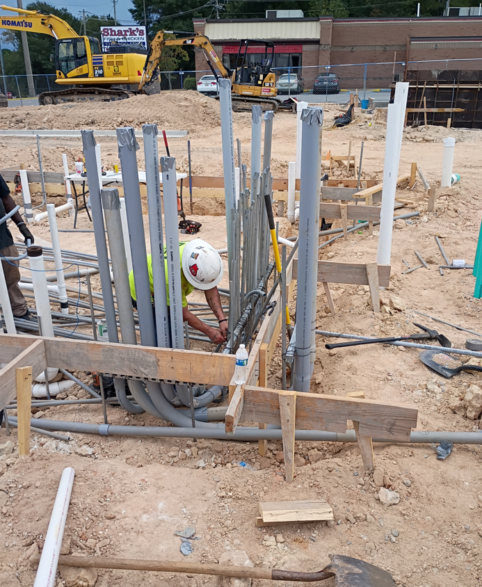 Construction site with workers, rebar, and pipes around a concrete foundation excavation