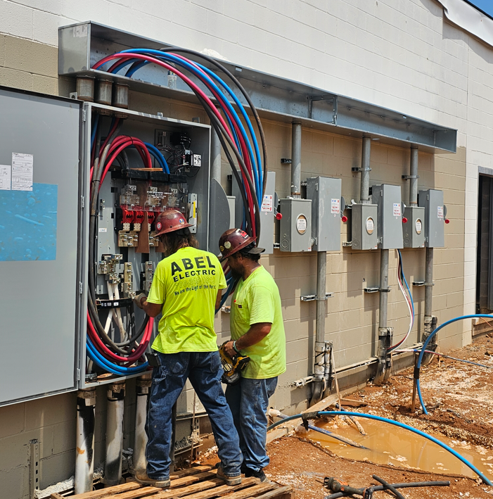 Two workers install electrical equipment and colored cables beside a building exterior.
