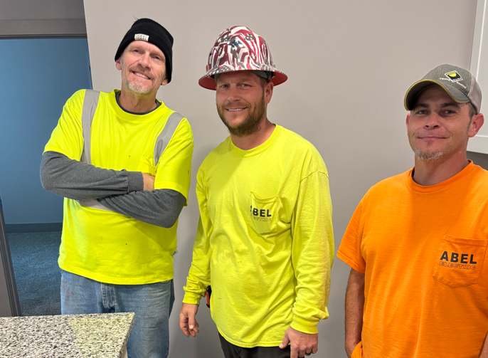 Three workers in neon safety shirts and hard hats stand together indoors, smiling.