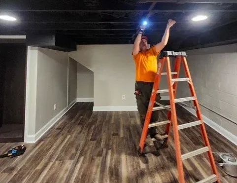 Worker on a ladder installing lights in a finished basement with wood-look flooring and gray walls.