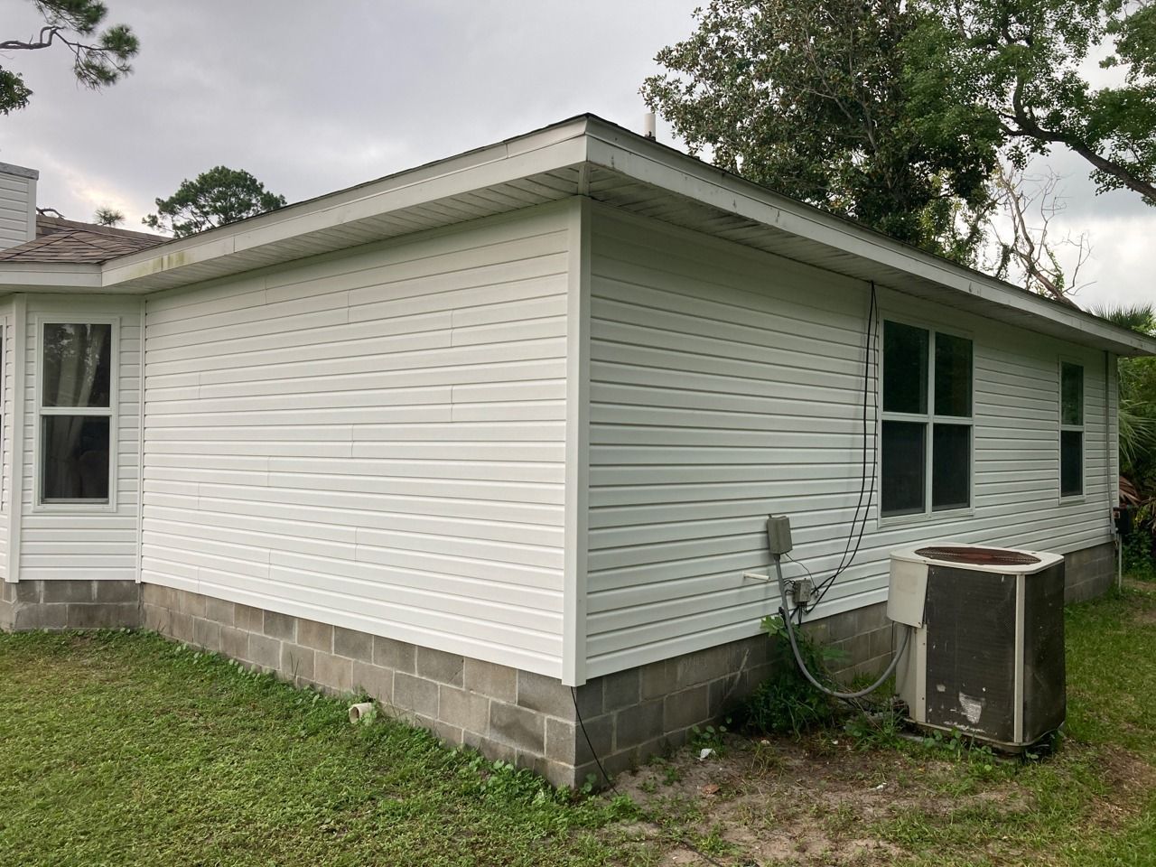 White siding on the corner of a house with visible concrete block foundation and an AC unit on the grass.