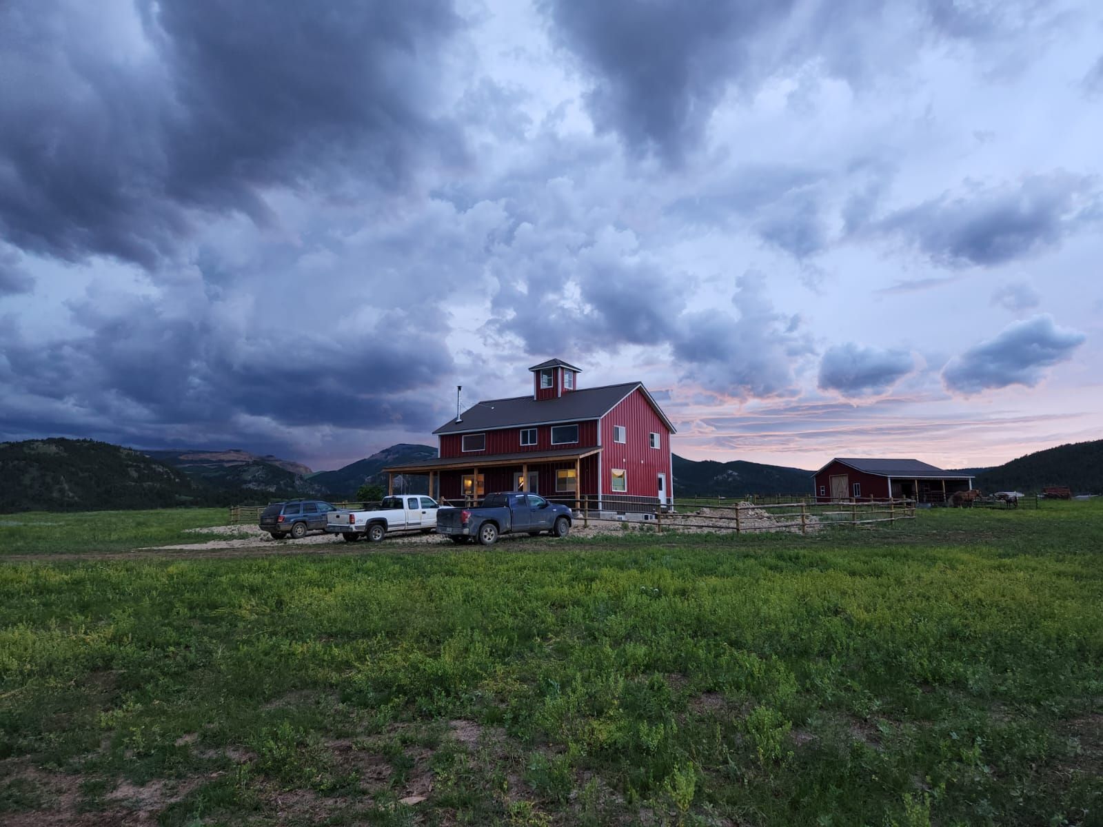A red barn is sitting in the middle of a grassy field with cars parked in front of it.