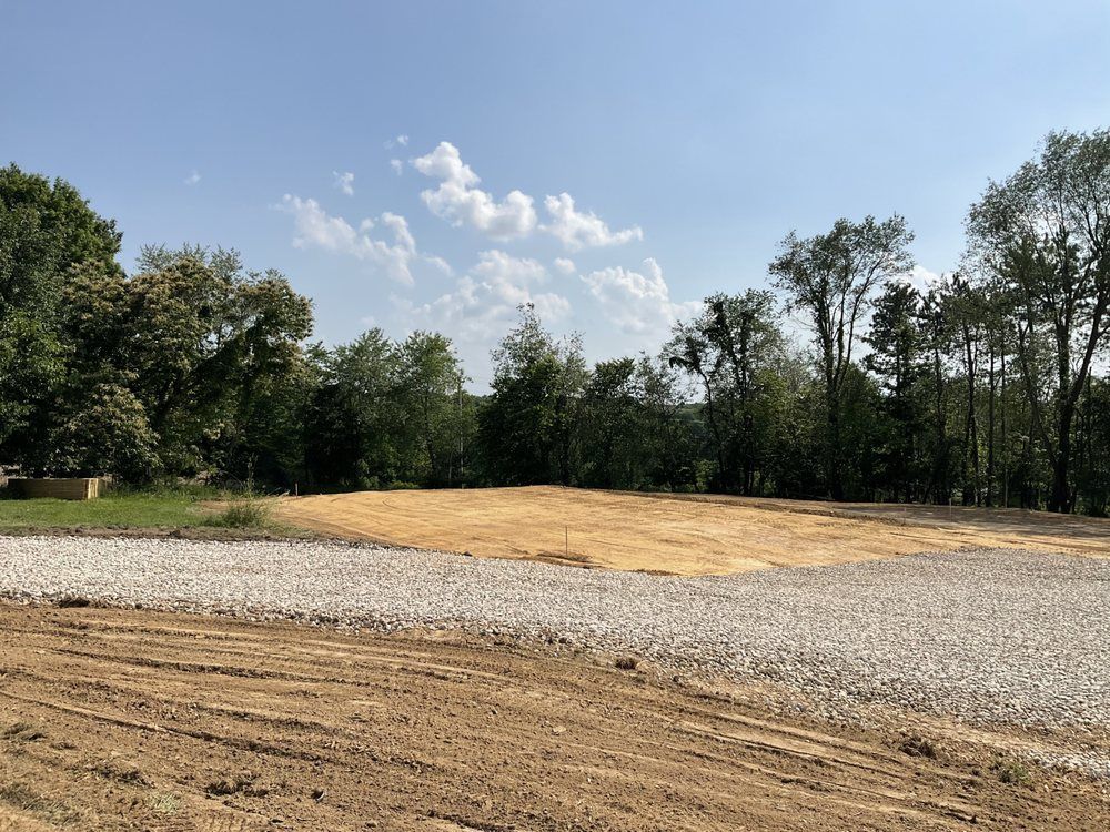 Gravel lot beside a dirt clearing and trees under a blue sky with scattered clouds