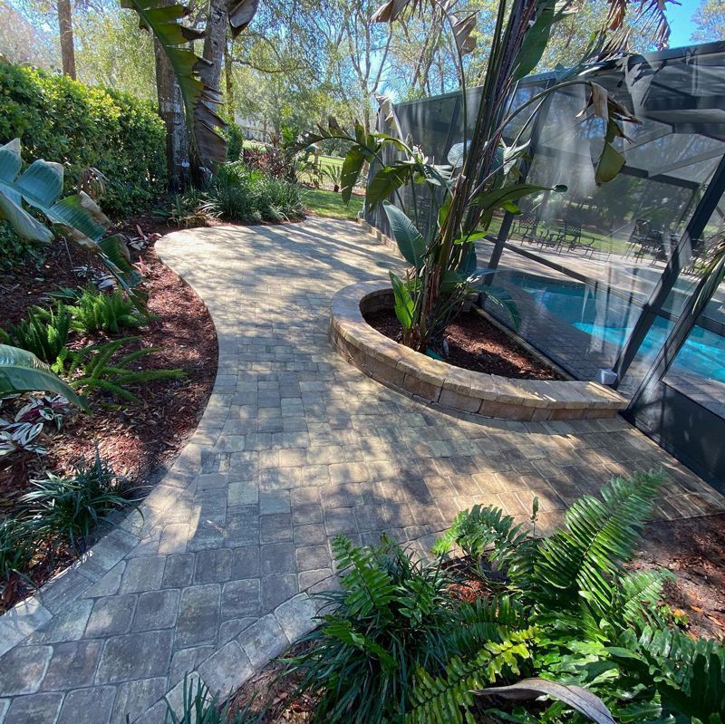 A brick pavement leading to a swimming pool surrounded by plants and trees.pa