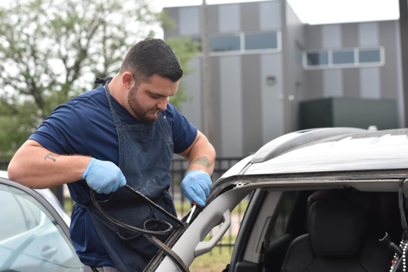 A man in blue gloves works on a car window. He's outside, wearing an apron, near a building.