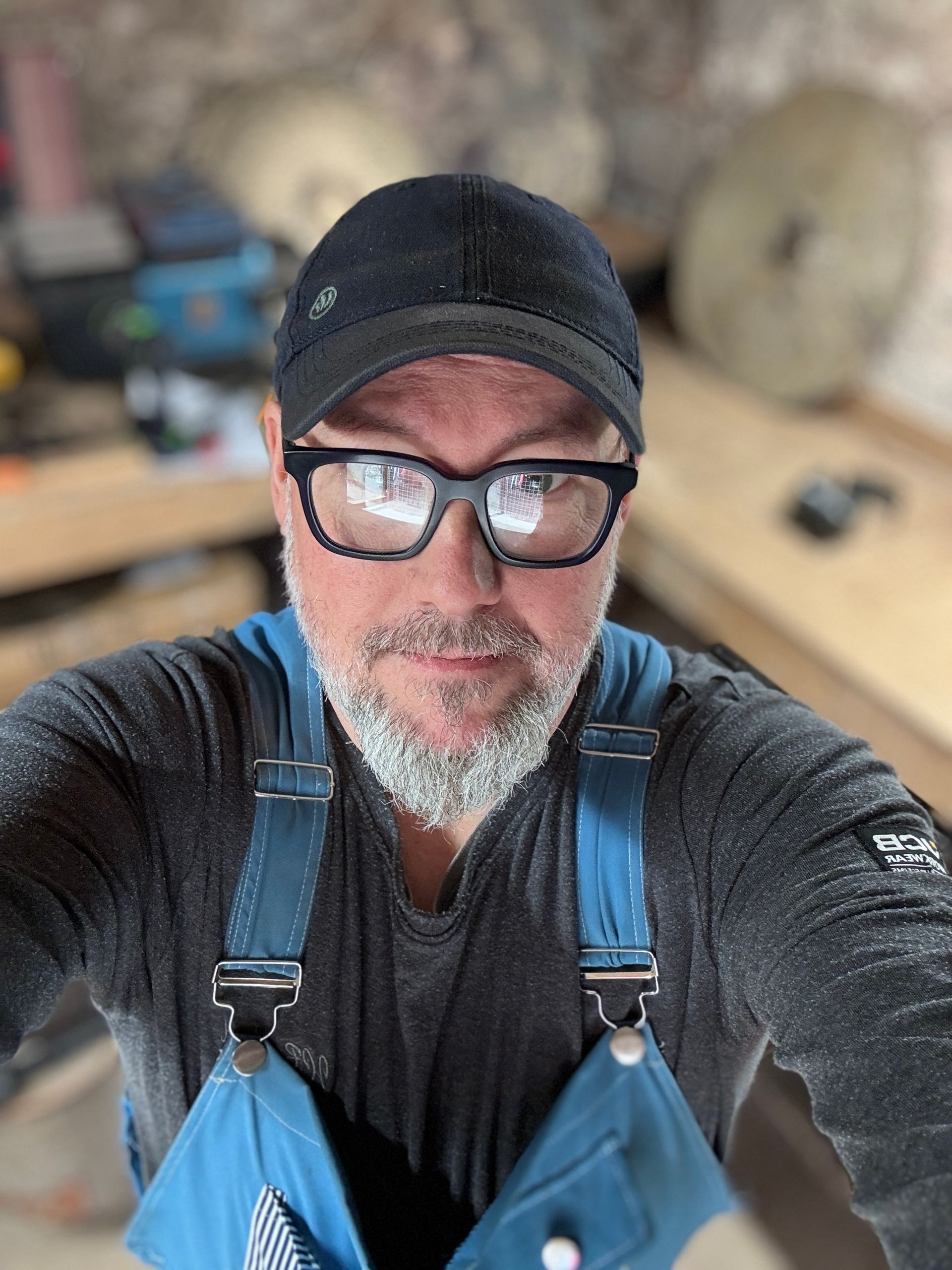 Man in a workshop wearing a black cap, glasses, and blue overalls, covered in sawdust.