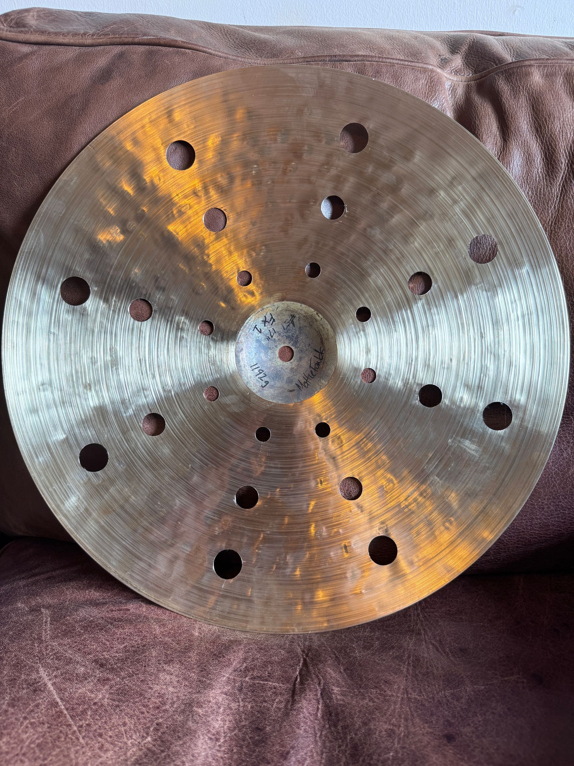 A bronze-colored drum cymbal with numerous evenly spaced holes, resting on a brown surface.