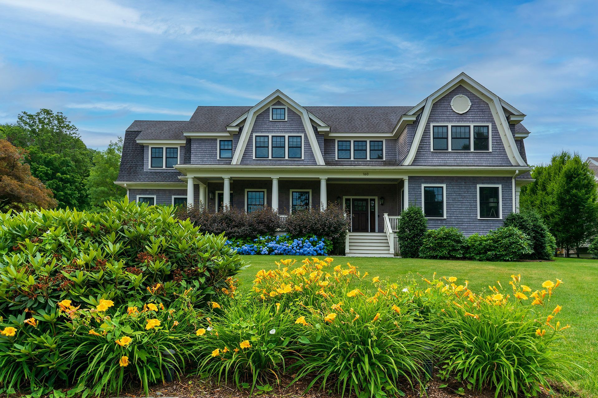 A large gray house with a large lawn and flowers in front of it.
