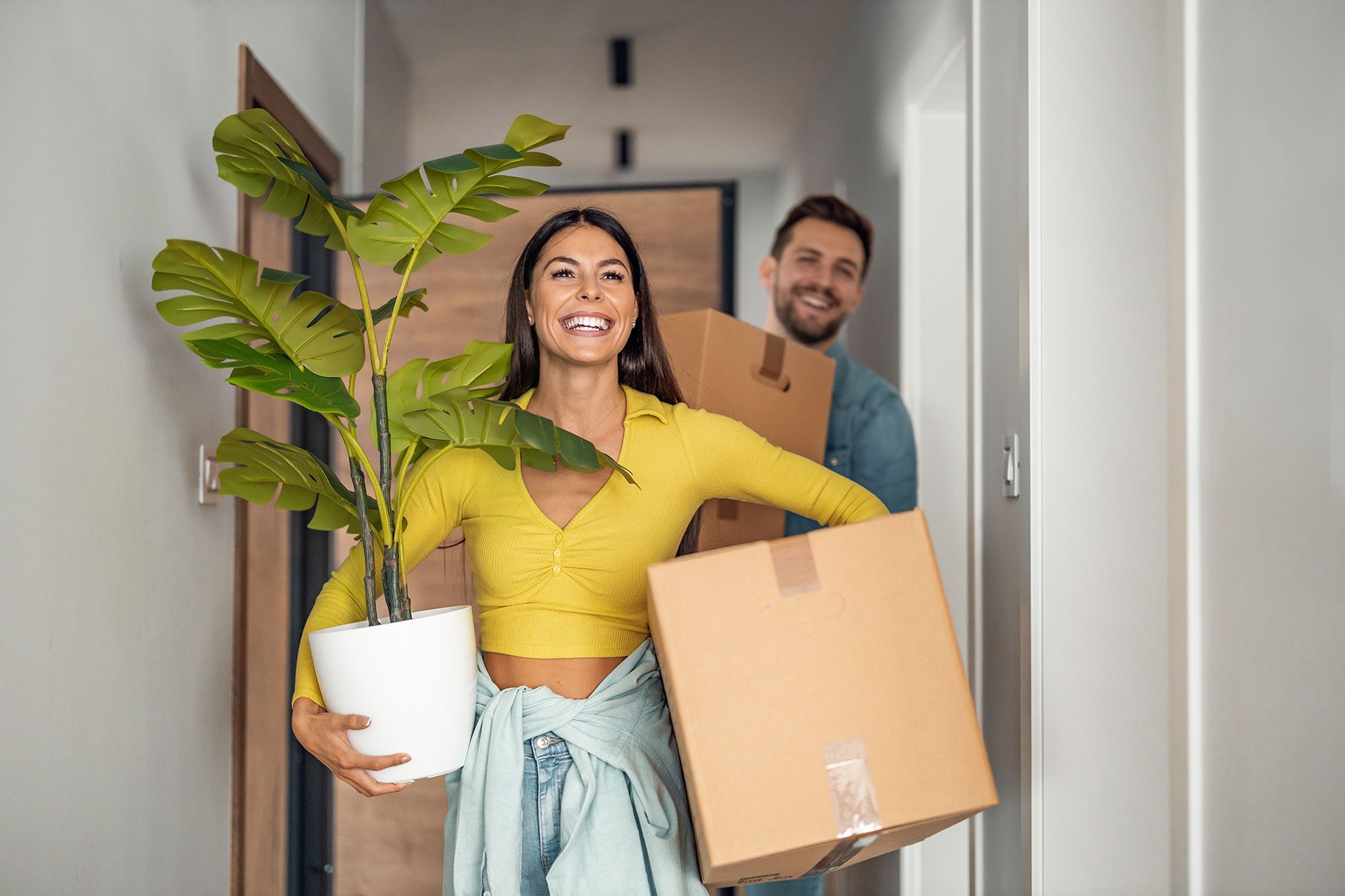 A man and a woman are carrying boxes and a potted plant into their new home.