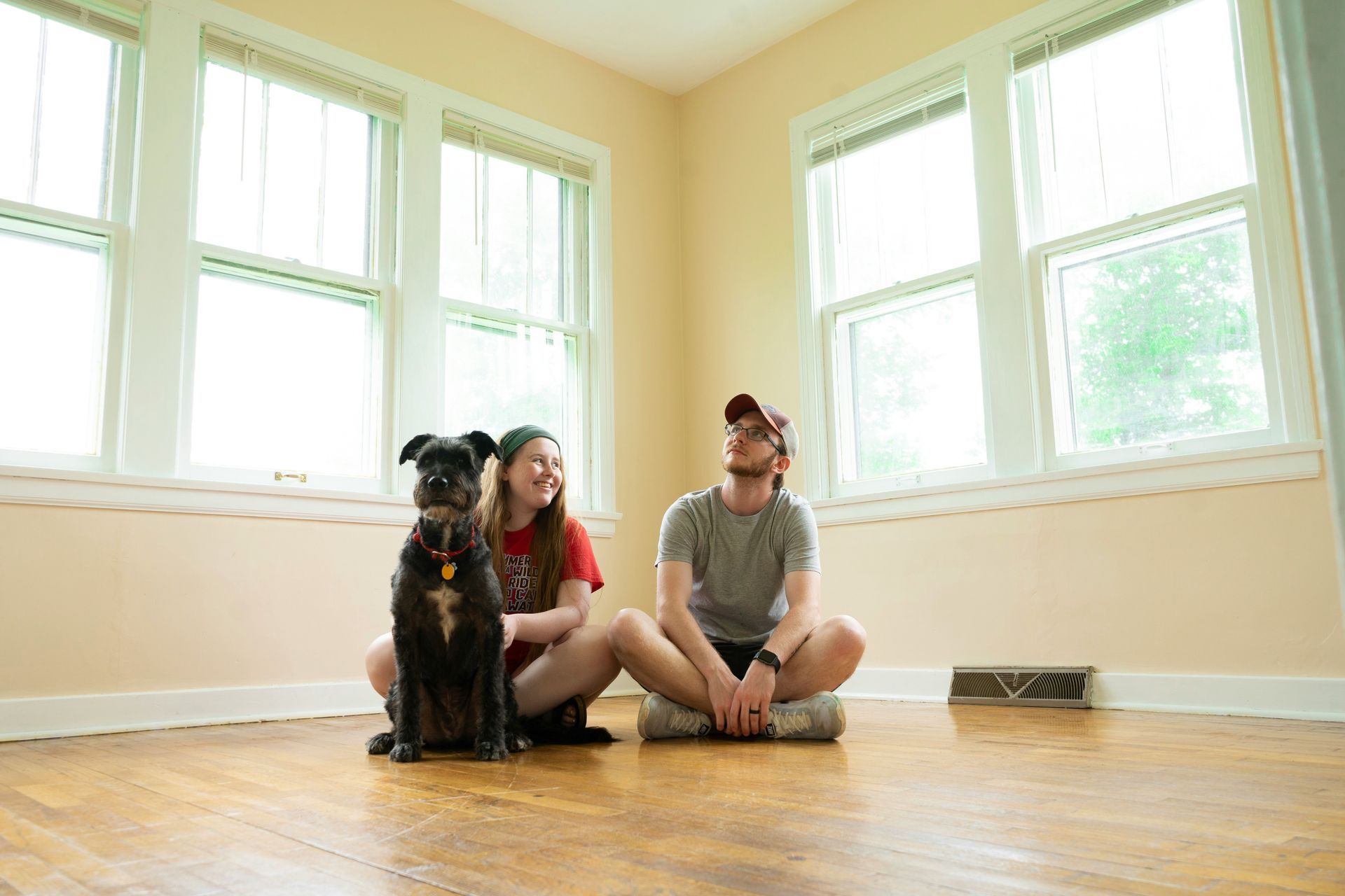 Couple and dog sitting on a hardwood floor in an empty room, looking up toward the windows.