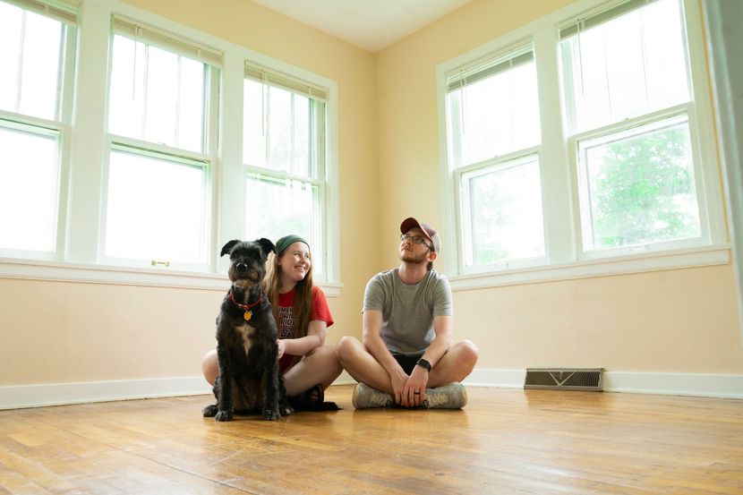 Couple and dog sitting on a hardwood floor in an empty room, looking up toward the windows.