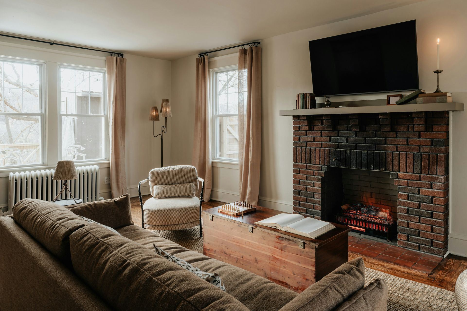 Cozy living room with brick fireplace, large TV, brown sofa, and windows with light curtains.