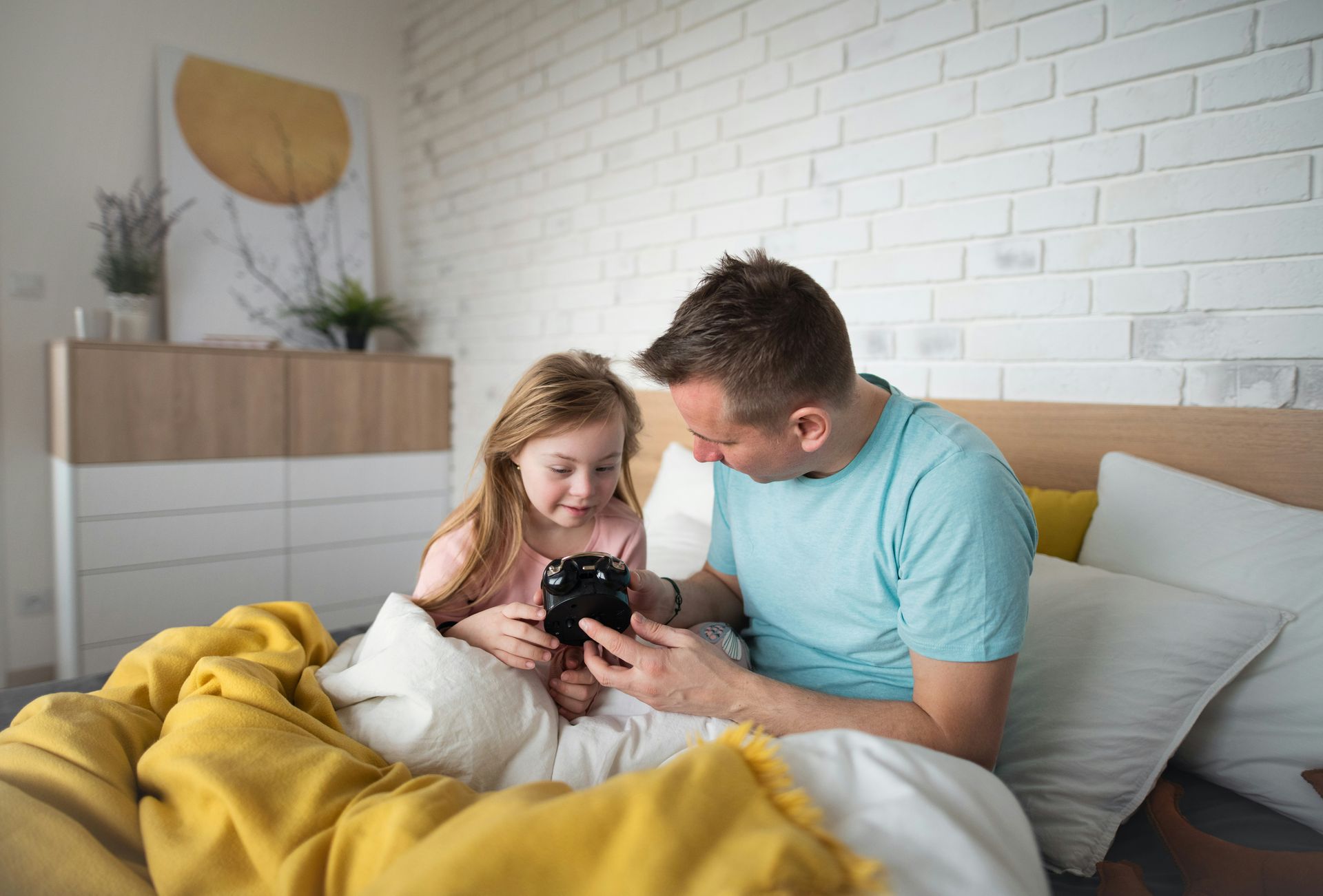 Man and girl in bed looking at a black clock. Yellow blanket, white brick wall.