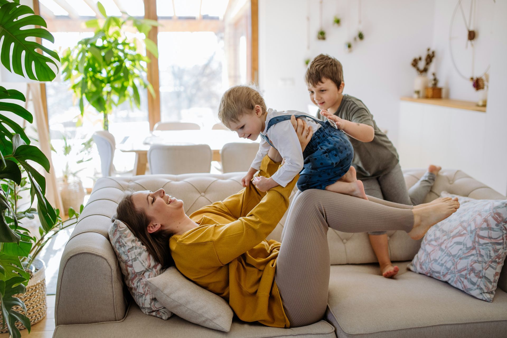 Woman lying on a sofa playfully holds a child as another child watches, bright interior.