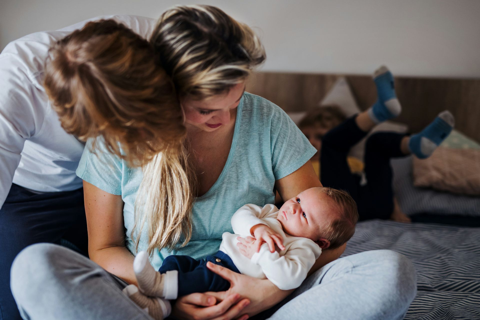 Family in bedroom, mother holding newborn. Father leans in, child's feet in background.