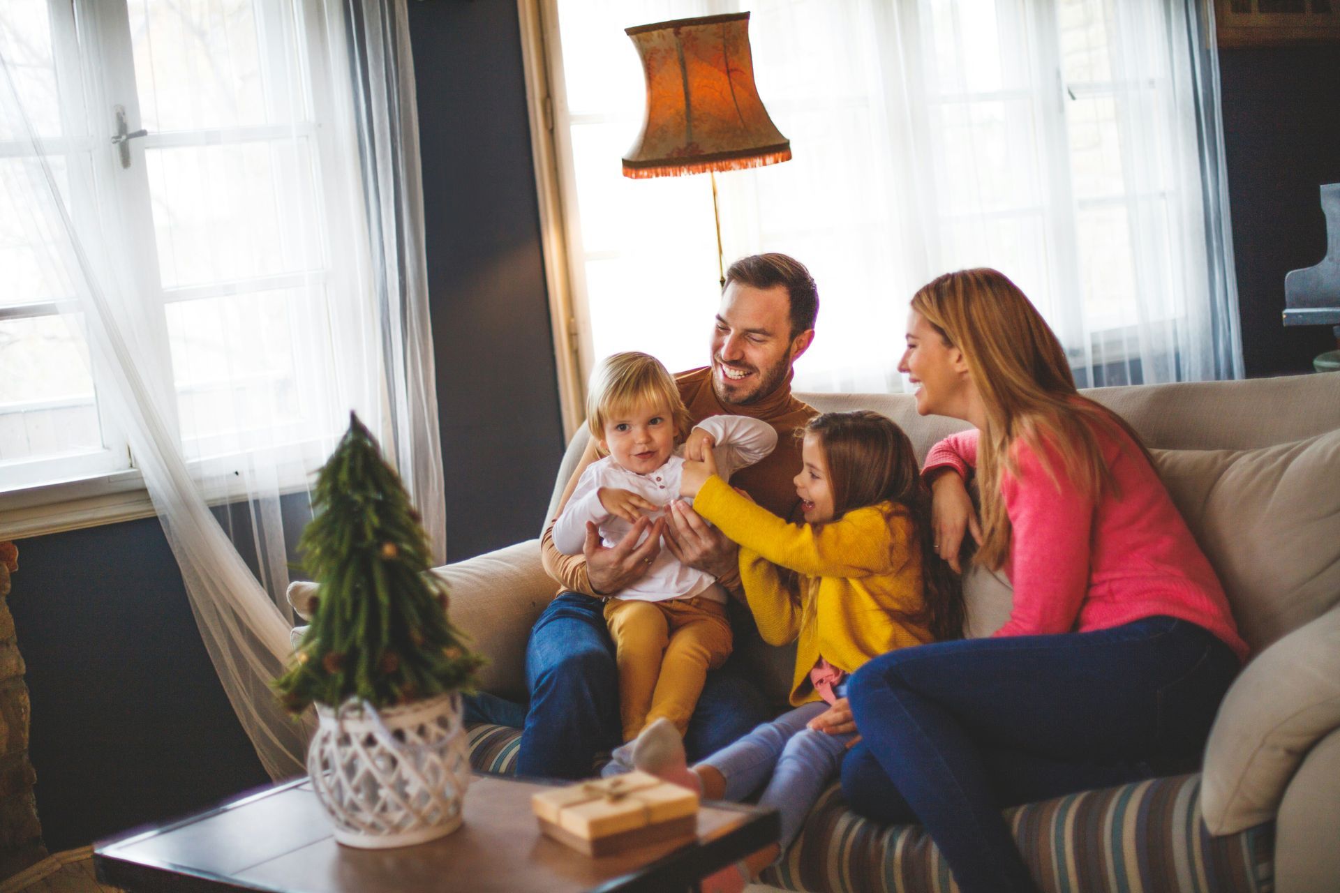 Family of four seated on a sofa, laughing. Two young children, and parents in a cozy living room.