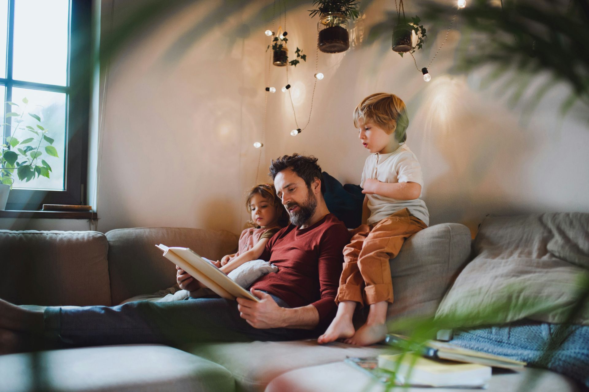 Man reading to two children on a sofa, cozy living room, string lights, book.