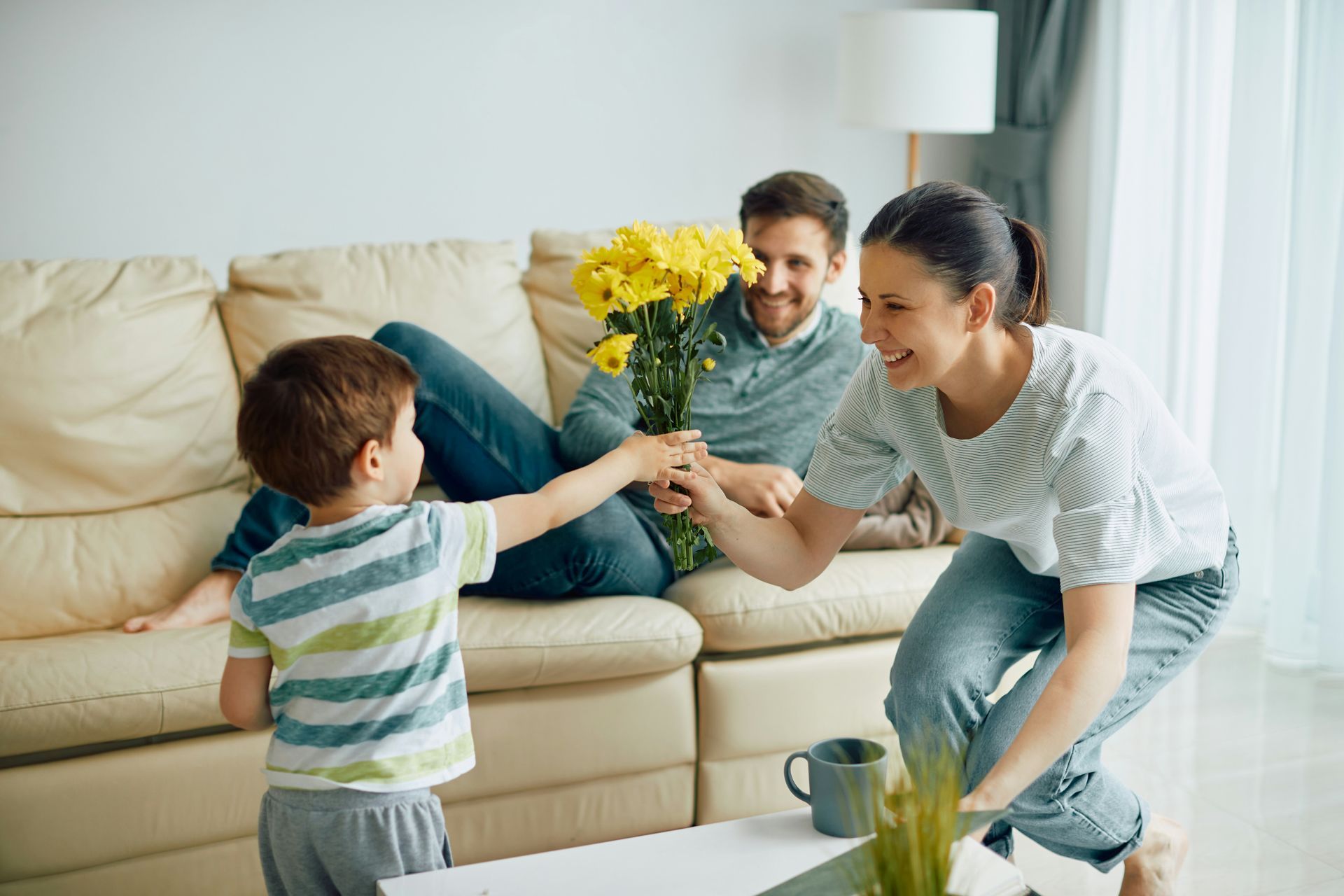 Child giving yellow flowers to smiling woman, man on couch. Modern living room.