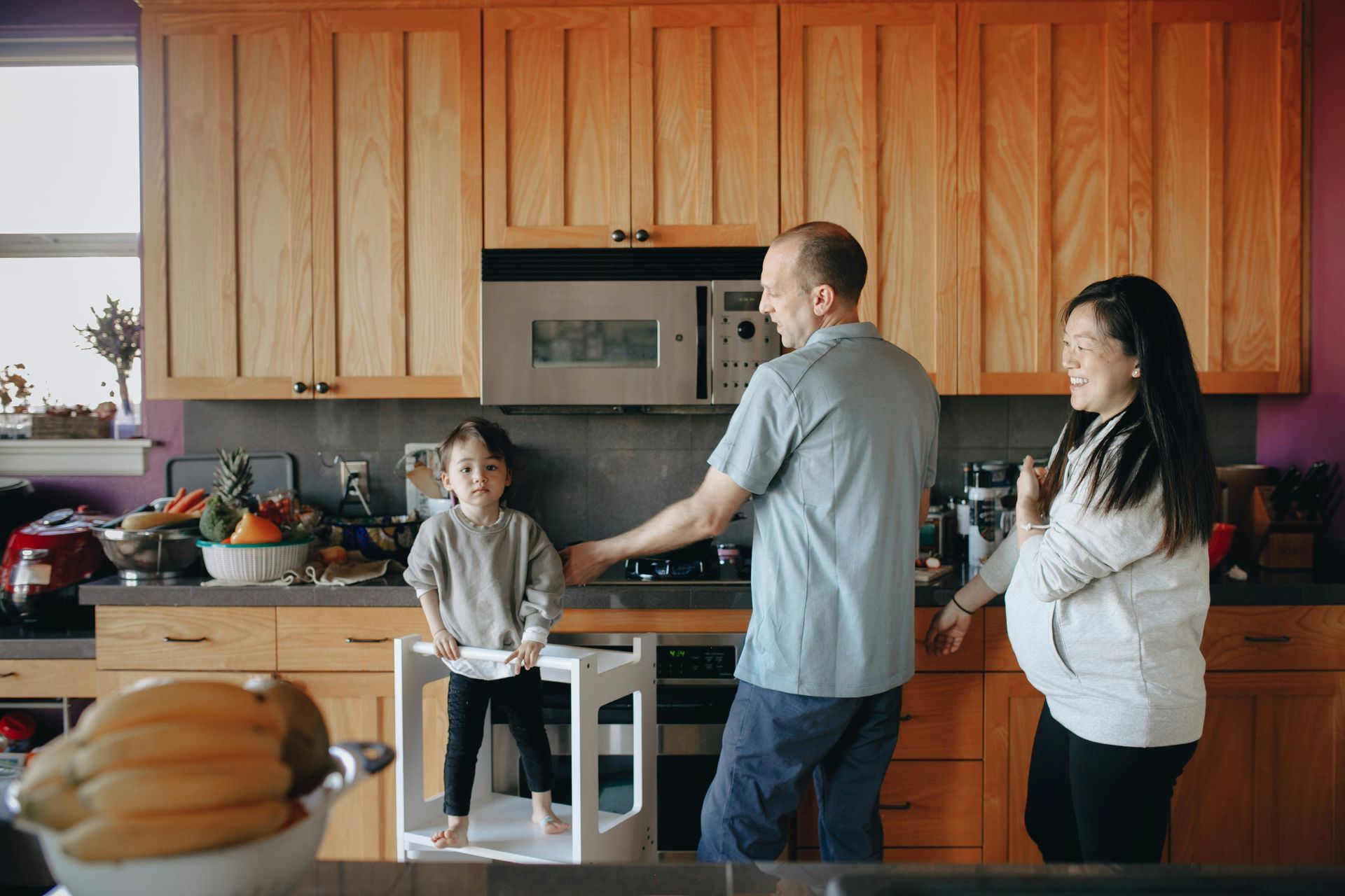 Family in kitchen: Child on step stool, parents near stove, smiling. Light wood cabinets, purple wall.