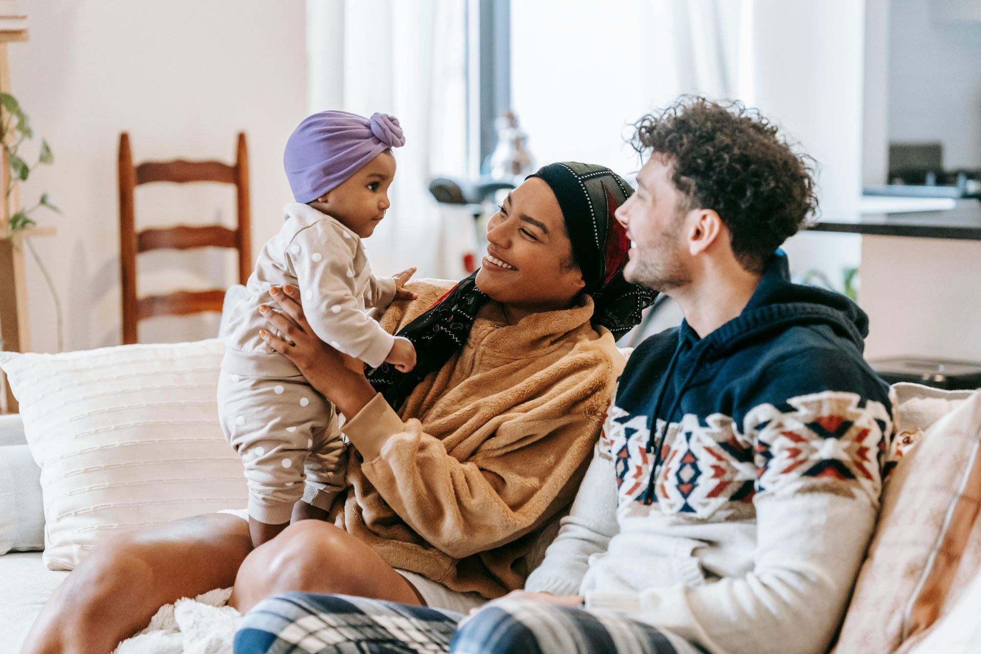 Couple on couch with baby, smiling. Woman holds baby wearing purple turban, warm-toned interior.