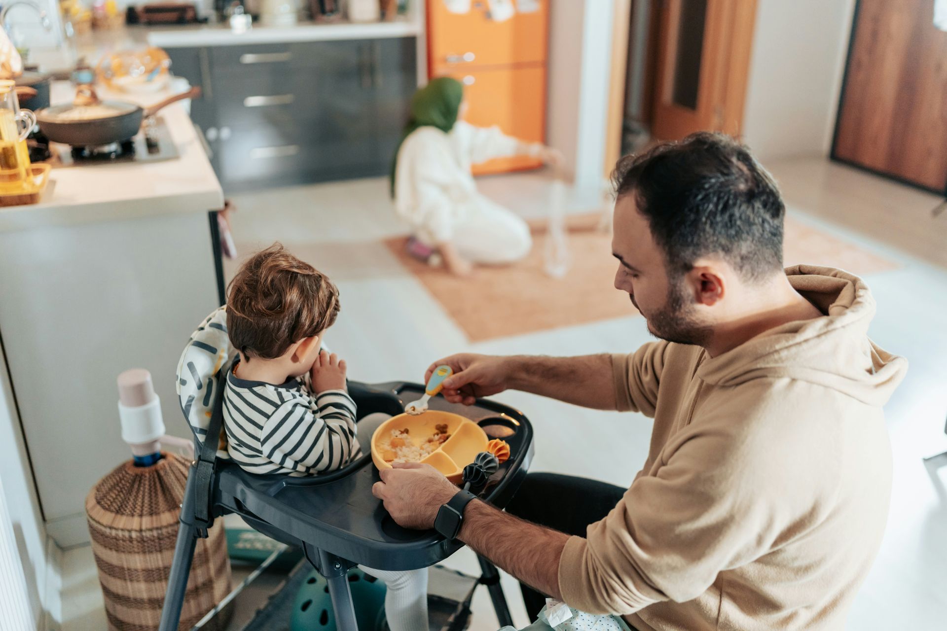 Man feeds a child in a highchair while a person prays in the background in a kitchen.