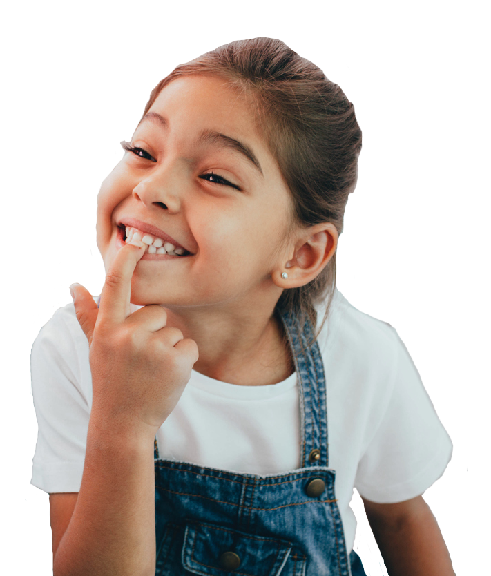 A little girl in overalls is smiling and holding her finger to her mouth.