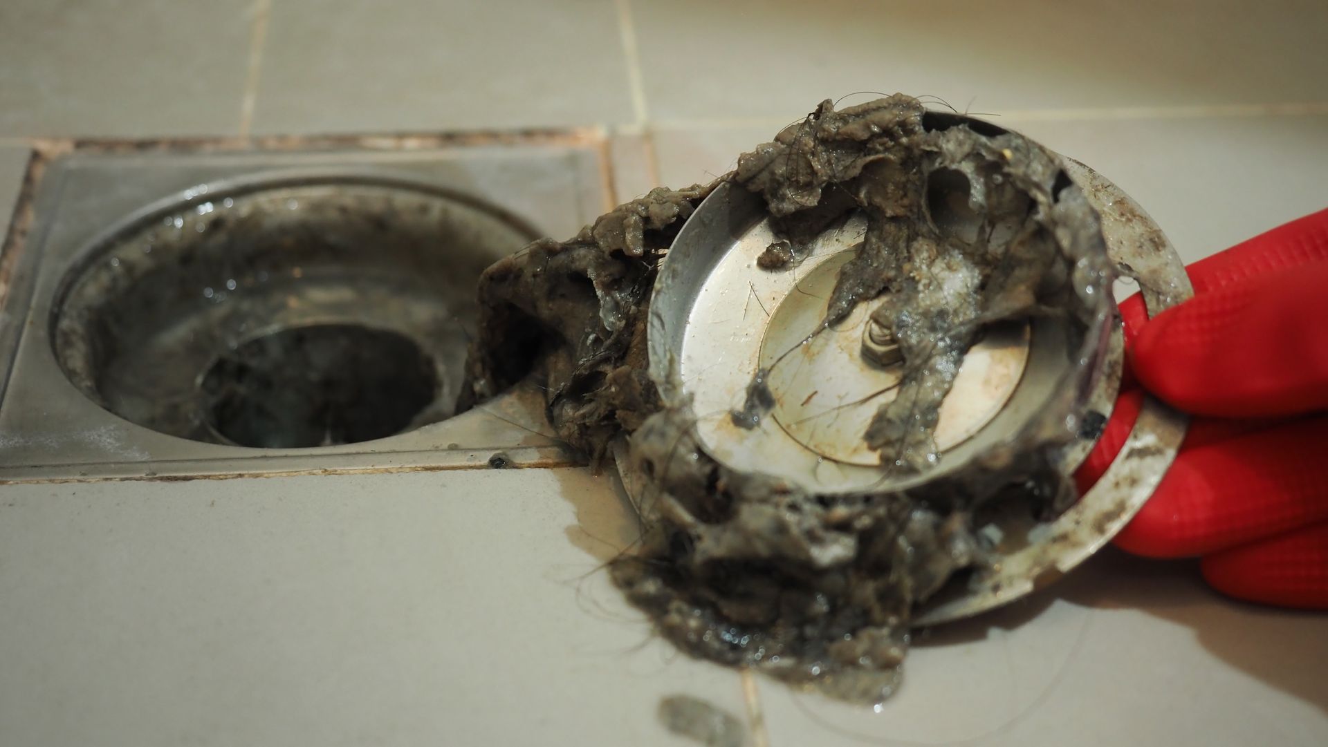 Dirty sink drain cap held by a red-gloved hand, covered in grime and hair.