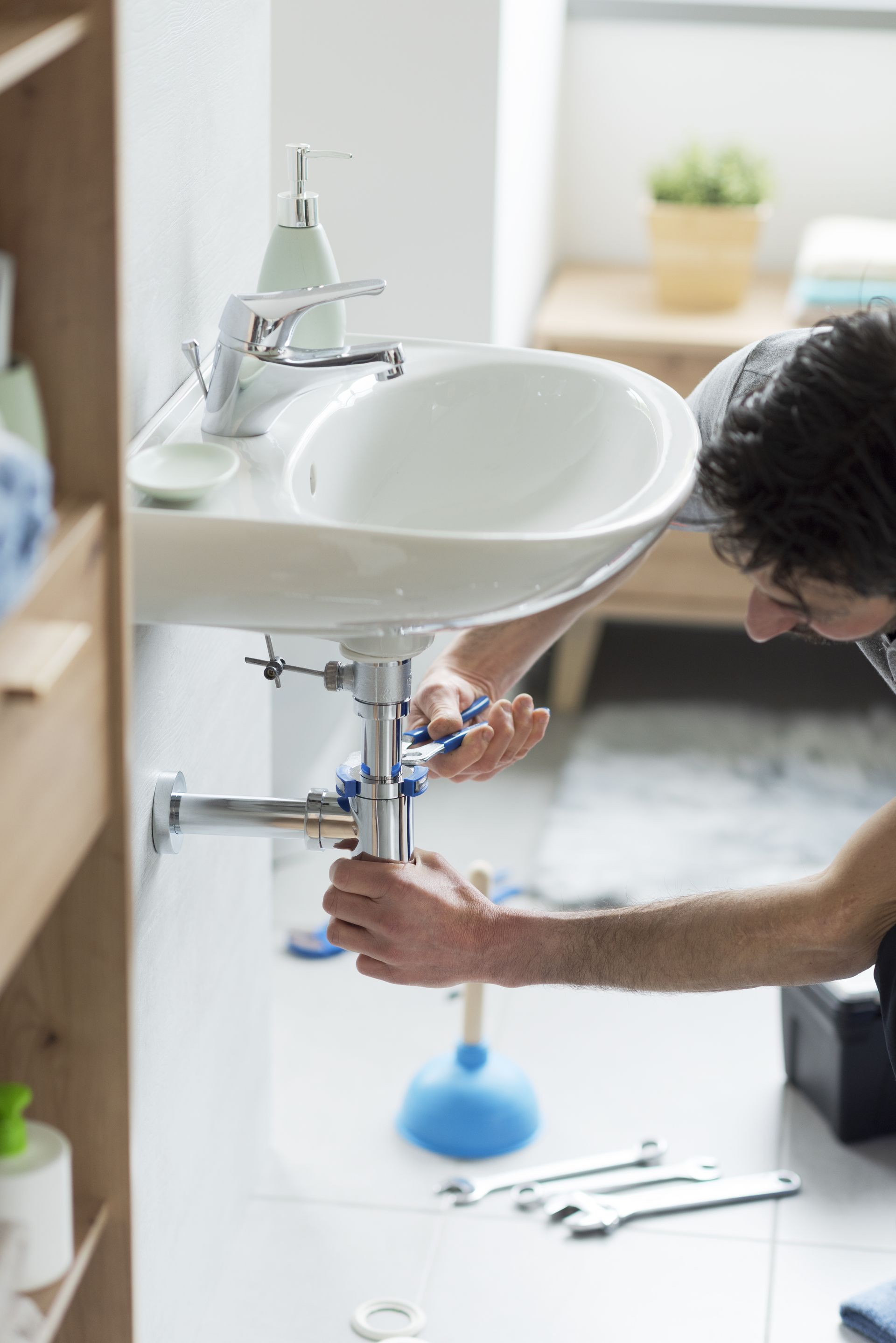 Person fixing plumbing under a white bathroom sink with tools on the floor
