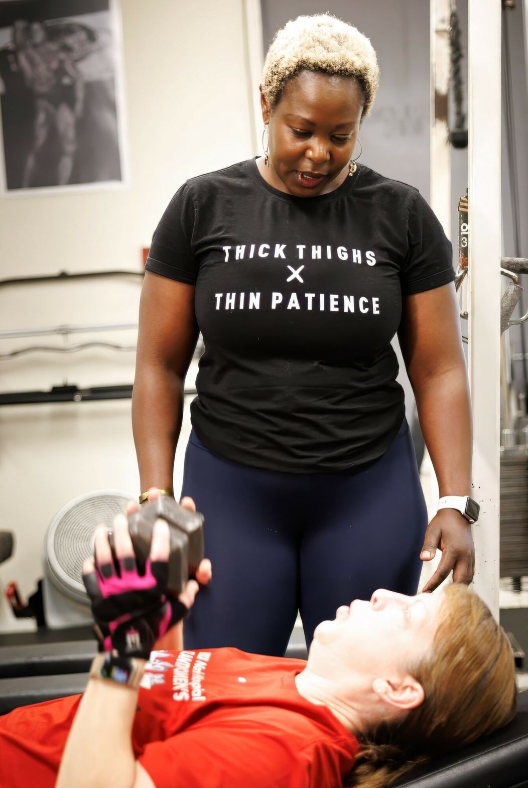 Woman assisting another in weightlifting at a gym. Woman in black shirt with text, other in red.