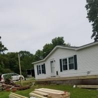 A white modular home with dark shutters sits in a yard with construction materials and a white car in the background.