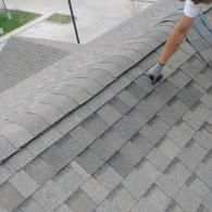 A person's gloved hand lifts a loose, curled shingle on a gray residential roof.