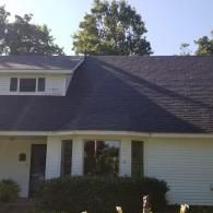 A single-story white house with a dark shingled roof, a front bay window, and a door partially hidden by a bush.