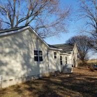Light-colored vinyl-sided house with a shingled roof under a clear blue sky in a yard with bare trees.