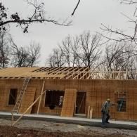 A person stands on the construction site of a wooden-framed building with a partially finished roof under bare trees.