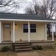 A single-story, light yellow house with a covered front porch, white pillars, a tan door, and a gray shingled roof.