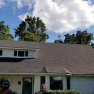 A residential house with a dark gray shingled roof, white siding, a garage, and large trees against a blue, cloudy sky.
