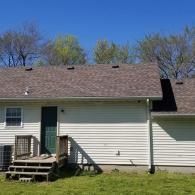 The back of a one-story house with cream siding, a shingled roof, a green door, and small wooden stairs on a grassy lawn.