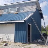 A blue-sided garage with a white door, new metal roof, and an extension ladder leaning against the side.