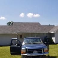 A dark Ford pickup truck parked in front of a ranch-style house under a sunny, partly cloudy blue sky.