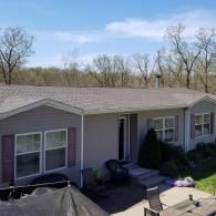 A gray manufactured home with a brown shingled roof, set against a backdrop of trees under a bright blue sky.