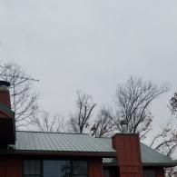 A metal roof on a wooden house with a red brick chimney under a cloudy sky with leafless trees in the background.