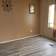 A room with light brown walls, wood-look flooring, a decorative metal wall hanging, a small framed picture, and a window.