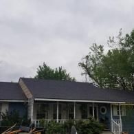A blue one-story house with a grey roof and a front porch, surrounded by green trees under a cloudy sky.