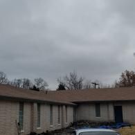 A low-angle view of a beige brick building with a brown shingled roof under a gray, cloudy sky.